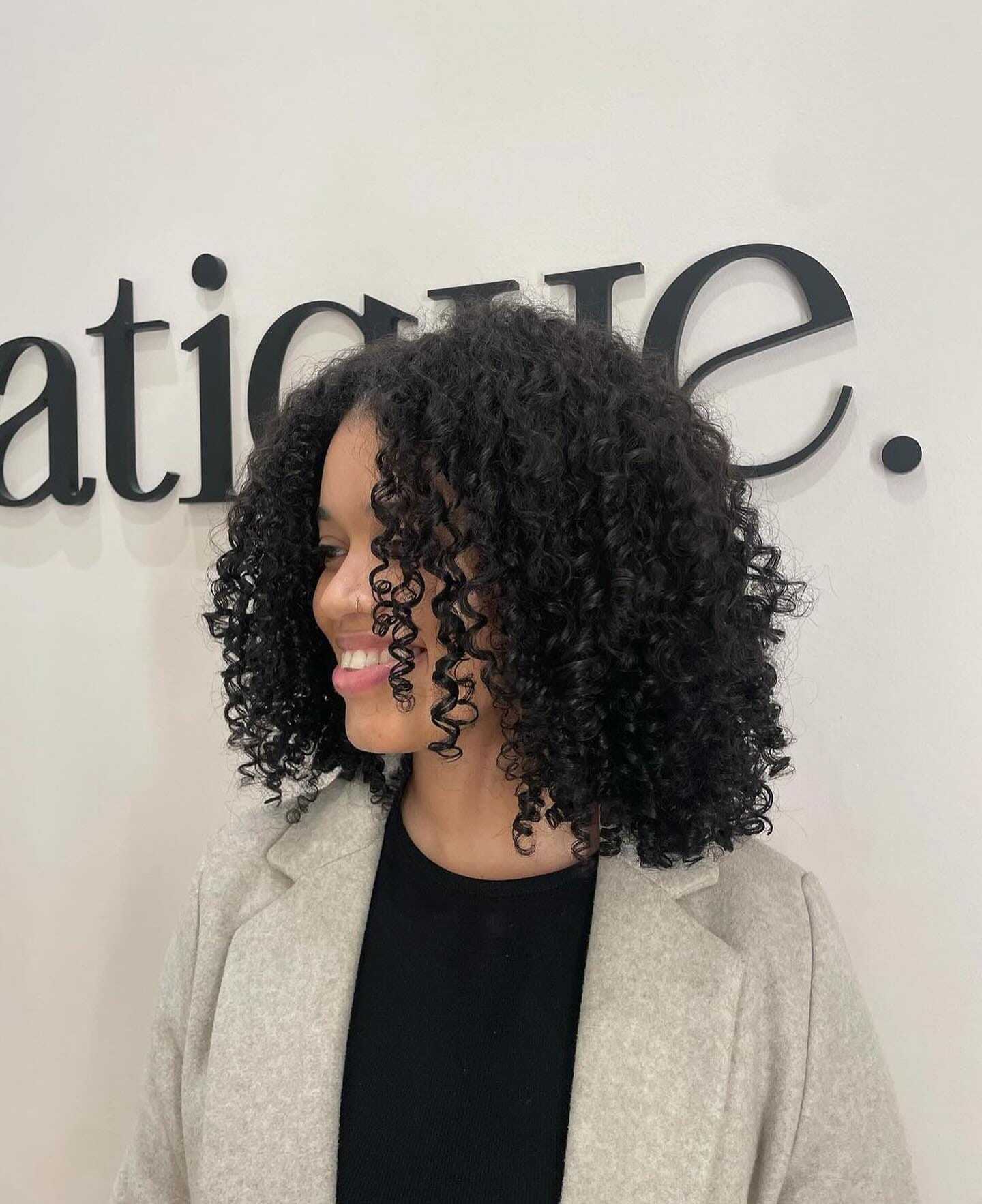 Smiling person with curly hair and beige coat in front of a wall with text.