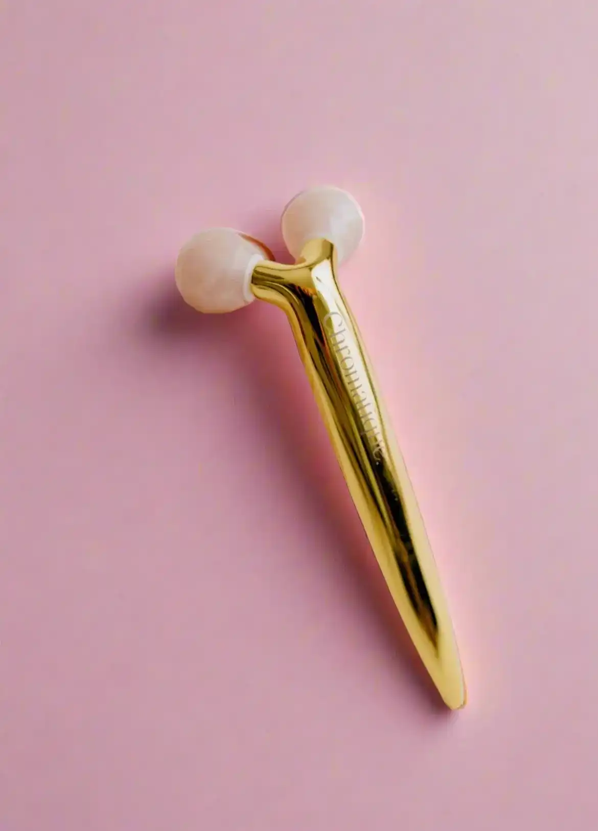 Gold facial roller with pink spheres on a pink background.