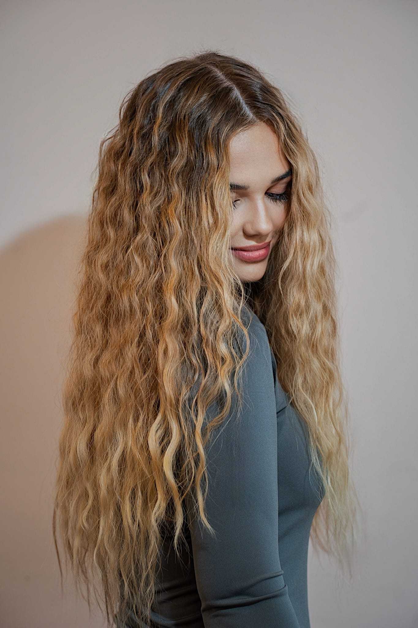 Young woman with long, wavy blonde hair, wearing a gray top, looking down and smiling.