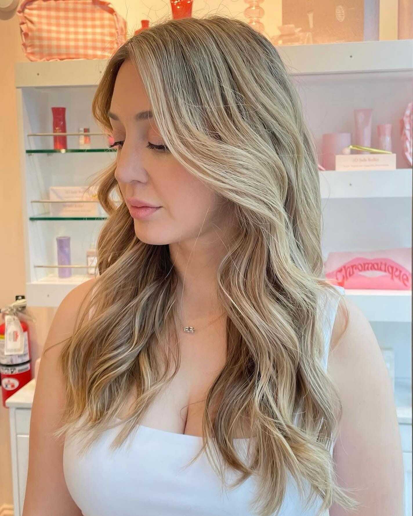 Woman with long, wavy blonde hair in a salon setting, wearing a white top.