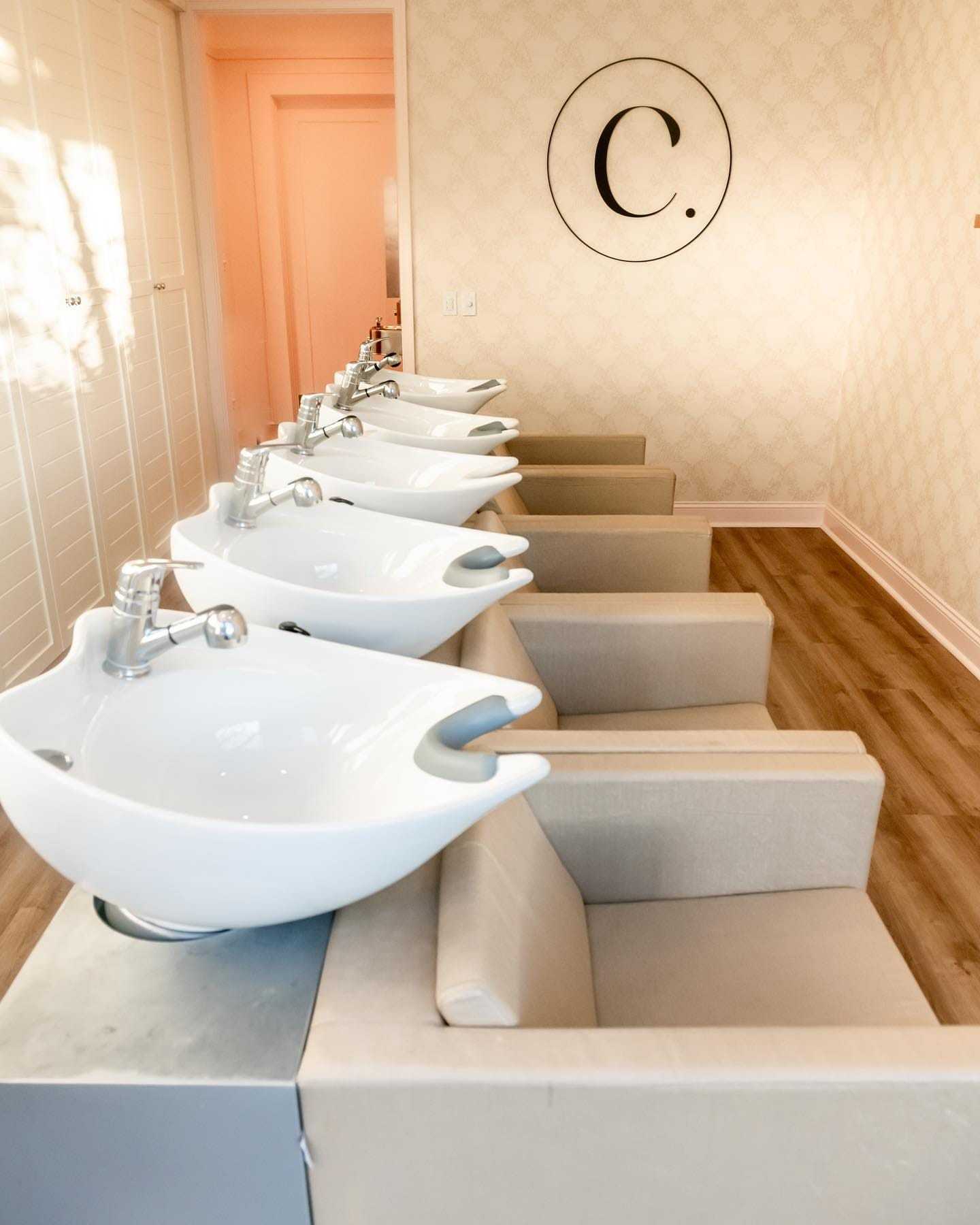 Modern hair salon with row of white sinks and beige chairs on wooden floor.