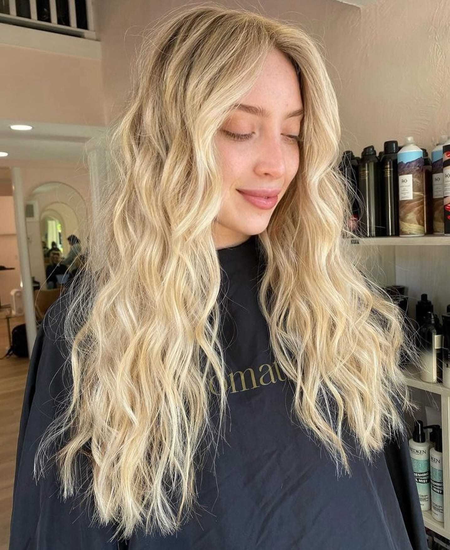 Long, wavy blonde hair styled in a salon with hair products visible in the background.