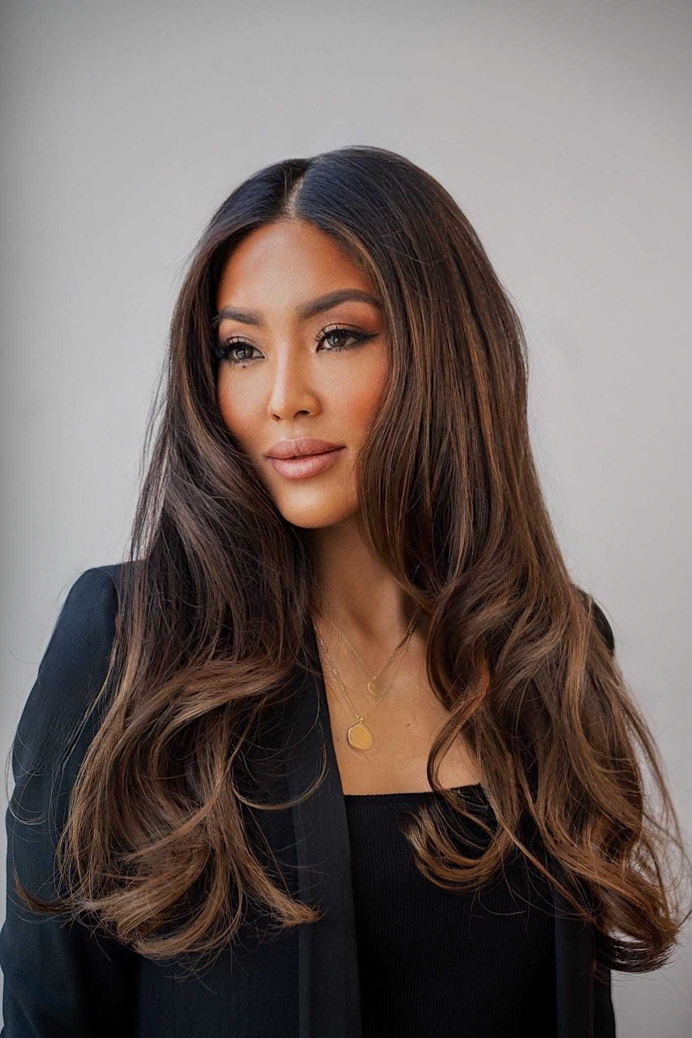 Woman with long, brown hair wearing a black suit and gold necklace against a neutral background.