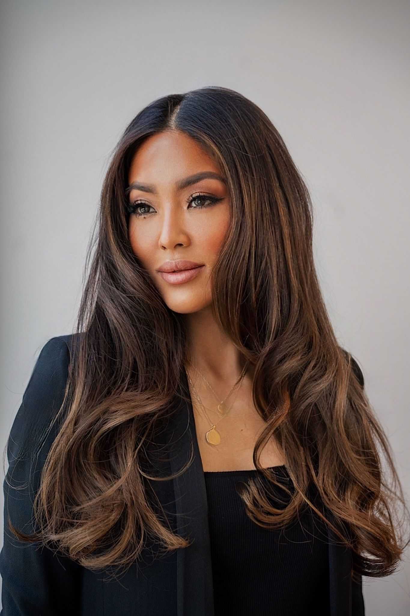 Woman with long, brown hair wearing a black suit and gold necklace against a neutral background.