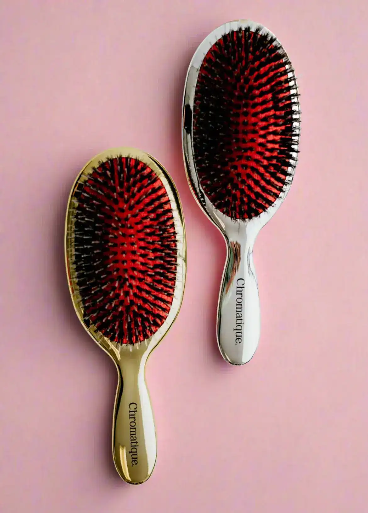 Two Chromatique hairbrushes with red bristles on a pink background.